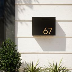 a house number sign mounted to the side of a white building with plants in front of it