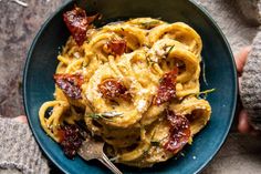 a blue bowl filled with pasta and bacon on top of a table next to a person's hand