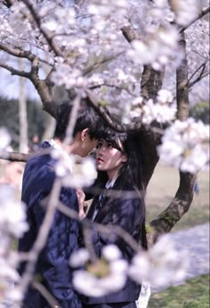 a man and woman standing next to each other under a tree with white flowers on it