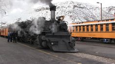 an old fashioned steam engine train traveling down the tracks near snow covered mountains and people