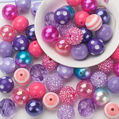 a white bowl filled with lots of purple and pink beads next to other colorful balls