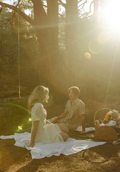 a man and woman sitting on a blanket in the woods, talking to each other