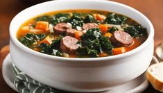 a bowl of soup with sausage, kale and carrots next to some slices of bread