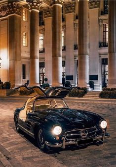 an old black mercedes sports car parked in front of a large building at night with lights on