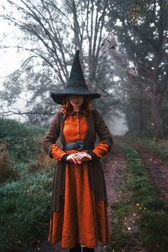 a woman dressed in an orange dress and black hat is standing on a path wearing a witch costume