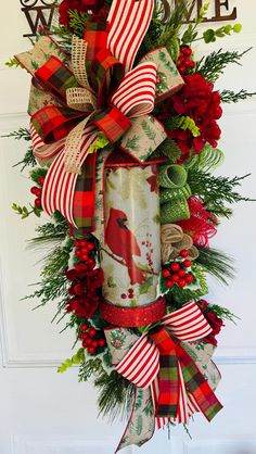 a christmas wreath hanging on the front door with red and green ribbons, evergreens, pine cones, holly berries, and a cardinal