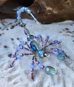 a beaded snowflake with blue and purple beads sits on the sand next to a rock