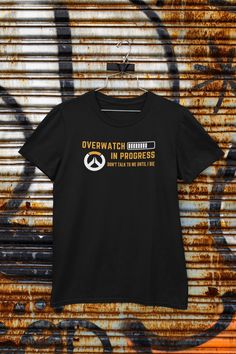 "Gamers, show off your dedication to Overwatch with our latest T-shirt design - the Overwatch In Progress T-Shirt! Made from 100% ring-spun cotton, this T-shirt is not only stylish but comfortable to wear. The front of the shirt features a bold \"Overwatch In Progress\" statement, letting everyone know that you're in the middle of a game and can't be bothered. The classic fit design ensures that you'll stay comfortable, whether you're playing for hours on end or just hanging out with your team. And with our high-quality construction, you can be confident that this T-shirt will last through all your intense matches. But that's not all, we've gone the extra mile to ensure our T-shirt is of the highest quality. With a seamless double-needle collar, fitted sleeves, and taped neck and shoulders Black Gamer T-shirt With Letter Print, Black Gamer T-shirt For Streetwear, Black Short Sleeve Gamer Tops, Black Gamer T-shirt With Screen Print, Black Gamer T-shirt With Graphic Print, Black Gamer Top With Screen Print, Black Short Sleeve Gamer T-shirt, Gamer Style Crew Neck T-shirt For Streetwear, Black Gamer Tops With Logo Print