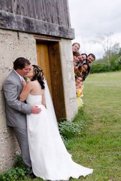 a bride and groom kissing in front of an outhouse with their wedding party behind them