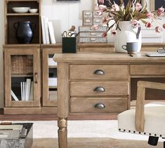 a wooden desk topped with a white vase filled with flowers next to a book shelf