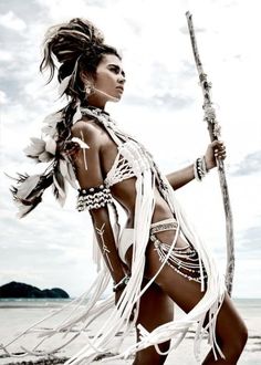 a woman in a white outfit holding a stick on the beach with water behind her