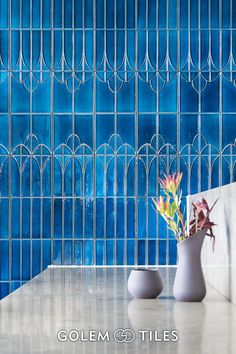 two vases with flowers sit on a table in front of a blue tiled wall