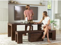 a woman sitting at a table with a man standing next to her in front of a flat screen tv