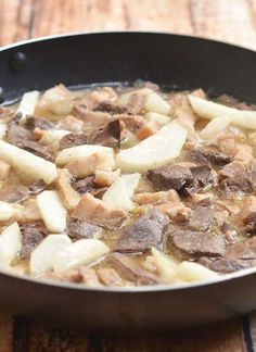 a skillet filled with meat and potatoes on top of a wooden table next to a knife