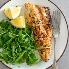 a white plate topped with fish next to a pile of greens and lemon wedges