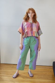 a woman standing in front of a white wall wearing green pants and a colorful shirt