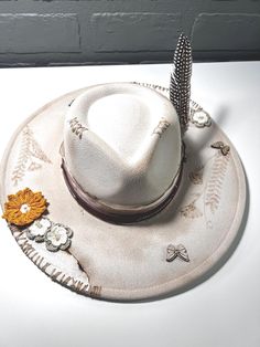 This wide-brim hat features a soft beige tone with intricate hand-stitched details along the crown and brim. Decorated with a mix of crochet flowers in warm earth tones and a single feather, it exudes a rustic, nature-inspired elegance. Hat Bands, Soft Beige, Oct 11, Wide Brimmed Hats, Hat Band, Brim Hat, Wide Brimmed, Earth Tones, The Crown