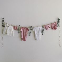 three baby ones hanging on a clothes line with flowers and plants next to them in front of a white wall