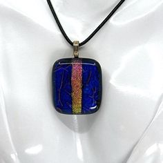 a necklace with a blue and yellow square shaped pendant on a black cord, hanging from a white mannequin