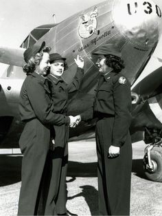 two women shaking hands in front of an old fashioned airplane with another woman standing next to it