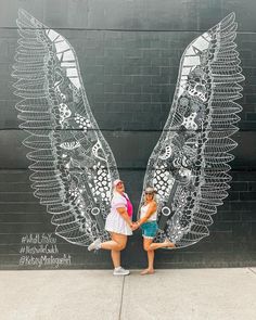 two women standing next to each other in front of a wall with wings drawn on it