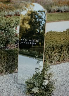 a mirror sitting on the side of a road next to bushes and flowers in front of it