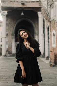 a woman standing in front of an old building wearing a black dress and holding her hands on her chest