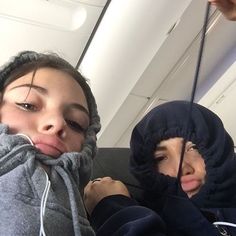 two women wearing hoodies on an airplane with their faces covered by sweats and pulling noses