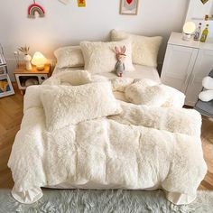 a bed covered in fluffy white blankets and pillows next to a night stand with stuffed animals on it