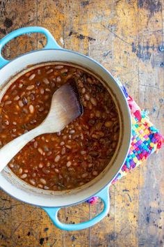 a pot filled with beans and a wooden spoon on top of a colorful cloth next to a wood table