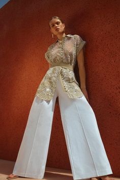 a woman standing in front of a red wall wearing white pants and a gold top