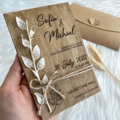 a hand holding a wooden wedding card with leaves and vines on it, next to a brown envelope