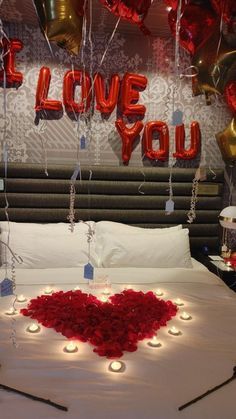 a bed topped with lots of red roses and candles next to balloons that spell i love you