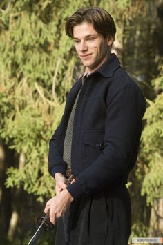 a young man in a coat and tie holding an umbrella while standing next to some trees