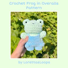 a hand holding a small crocheted frog toy in front of green plants and bushes