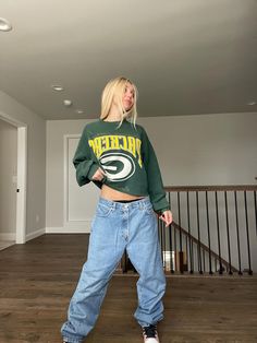 a woman standing on top of a wooden floor wearing jeans and a green bay packers sweatshirt