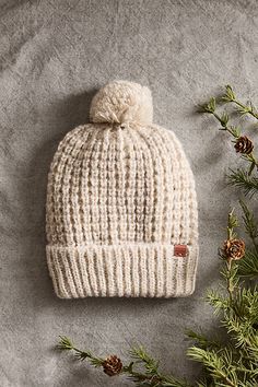 a white knitted hat with a pom - pom on the side next to pine branches