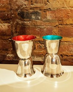 two silver cups sitting on top of a white table next to a brick wall and stone wall