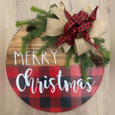 a wooden sign that says merry christmas with pine branches and plaid bow on it, sitting on a wood floor