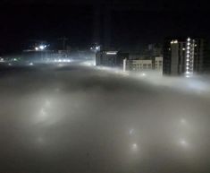 fog is covering the ground and buildings in the distance at night with lights shining on them