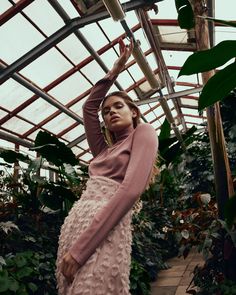 a woman in a pink sweater and skirt standing next to a plant filled greenhouse area