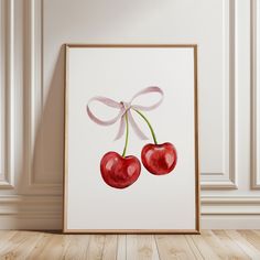 two cherries tied to a white ribbon in front of a framed art print on the wall