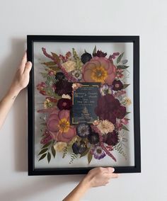 a person holding up a framed photo with flowers on the wall in front of them