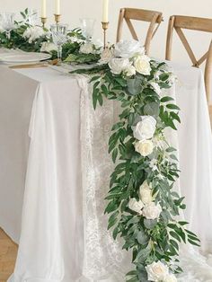 the table is set with white flowers and greenery on it, along with candles