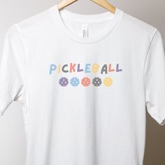 Introducing our new pickleball shirt, perfect for both on and off the court! Made from a lightweight fabric, this shirt will keep you comfortable no matter how long your pickleball game lasts. The relaxed fit ensures that you have full range of motion, allowing you to move freely and play your best. This shirt features a classic crew neck design and short sleeves, making it versatile enough to wear on any occasion. The shirt also features a fun and colorful design with a pickleball theme, showing off your love for the sport. The high-quality print will stay vibrant even after many washes, ensuring that you can enjoy wearing this shirt for years to come. This casual pickleball shirt is available in a range of sizes to fit every body type. It's perfect for both men and women and can be dress Fun White Sports T-shirt, Sporty White T-shirt For Pickleball, White Moisture-wicking Tennis T-shirt, White Moisture-wicking T-shirt For Tennis, White Tennis Top With Team Name, White T-shirt With Team Name For Pickleball, Pickleball Shirts, Pickleball Shirt, Pickleball Gift