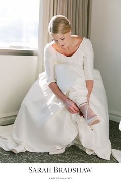 a woman in white dress sitting on the floor next to a window with her shoes off