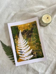 a card with a white fern leaf next to a lit candle on a bed sheet