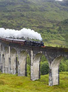 a train traveling over a bridge on top of a lush green hillside next to mountains