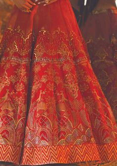 This dreamy lehenga features hand embroidery and gold printing. The blouse comes with a v neckline with floral motifs on it. A bridal wear lehenga that gives an exquisite look to you, bathed in wedding vibes. Festive Gown With Gold Embroidery For Traditional Ceremonies, Red Dola Silk Gown With Intricate Embroidery, Traditional Gold Embroidered Gown For Festive Season, Traditional Gown With Gold Embroidery, Red Dola Silk Gown With Resham Embroidery, Red Gown With Resham Embroidery In Dola Silk, Unstitched Gold Embroidered Sharara For Wedding, Designer Embroidered Brocade Lehenga, Elegant Designer Lehenga With Gold Embroidery