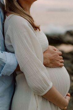 a pregnant woman and man standing next to each other near the ocean with their hands on her belly